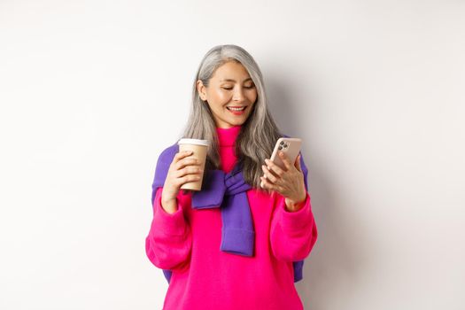 Trendy senior woman drinking coffee and using smartphone. Korean senior lady reading message on mobile phone and smiling, white background.