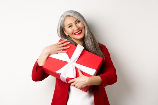 Happy asian grandmother hugging red gift box and smiling grateful, thanking for present, standing over white background.