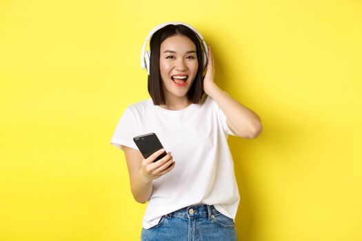 Cool asian girl dancing and listening music in wireless headphones, holding smartphone in hand, standing over yellow background.