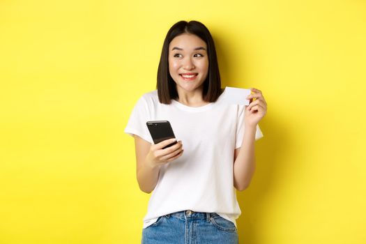 E-commerce and online shopping concept. Cheerful asian girl paying in internet, holding smartphone and plastic credit card, smiling and looking left, yellow background.