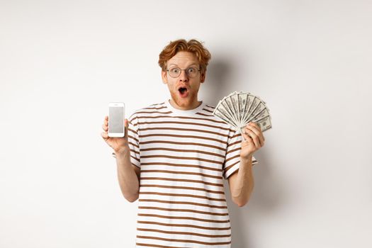 Amazed redhead man showing smartphone app on blank screen and money, winning prize cash online, standing over white background.