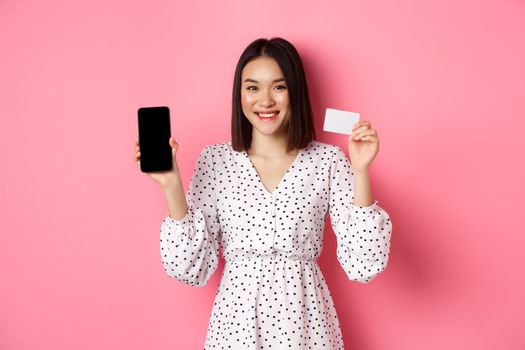 Cute asian woman shopping online, showing bank credit card and mobile screen, smiling and looking at camera, standing over pink background.