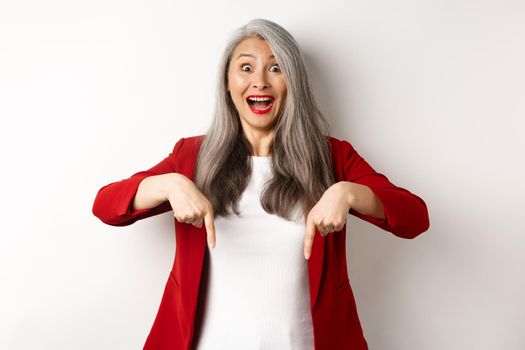 Portrait of happy asian lady in red blazer showing logo, pointing fingers down and smiling cheerful, check this out gesture, white background.