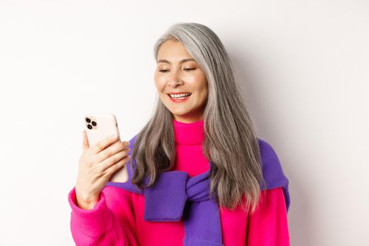 Close-up of fashionable asian senior woman looking at smartphone and reading message, smiling at mobile screen, standing over white background.