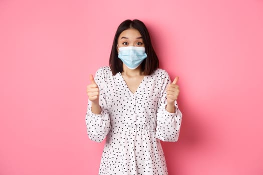 Covid-19, quarantine and lifestyle concept. Excited asian woman in face mask showing thumbs-up, praising good job, looking amazed at camera, standing over pink background.