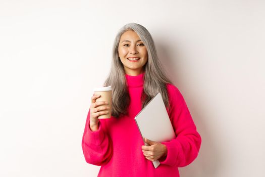 E-commerce concept. Happy asian senior freelancer, drinking coffee and holding laptop, smiling relaxed at camera, standing over white background.
