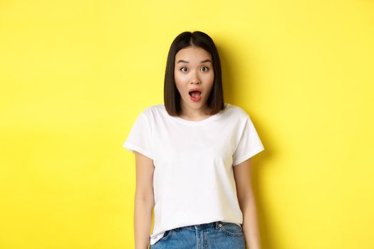 Beauty and fashion concept. Surprised asian girl open mouth and staring at camera, drop jaw impressed, standing over yellow background.