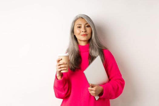 Stylish asian senior businesswoman drinking coffee and holding laptop, looking confident and smug at camera, standing over white background.