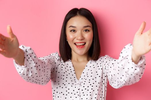 Close-up of cute asian woman reaching hands forward, taking selfie, record beauty blog and smiling, standing over pink background.