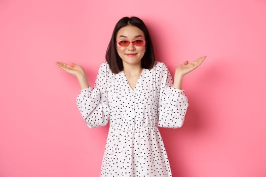 Cute asian woman tourist smiling at camera, shrugging clueless, dont know, wearing trendy sunglasses and white dress, standing against pink background.