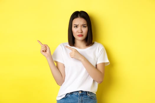 Beauty and fashion concept. Beautiful asian woman in white t-shirt pointing fingers left, standing over yellow background.