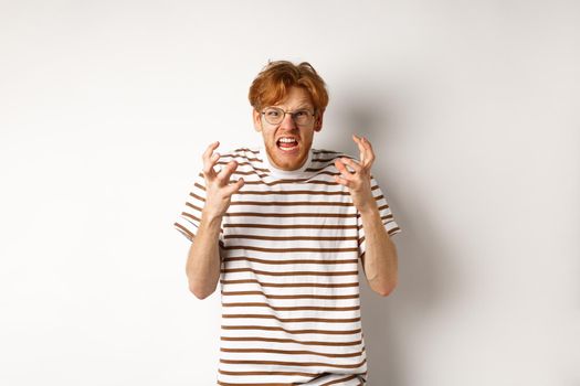 Portrait of redhead male student looking annoyed and pissed-off, shaking hands and staring angry at camera, shouting at someone, standing over white background.