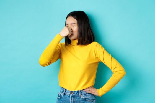 Image of asian stylish girl shut her nose and grimacing from aversion, something smell bad, standing disgusted against blue background.