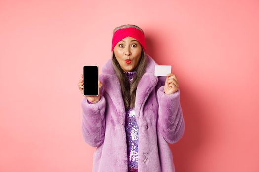 Special promotion. Excited asian senior woman showing blank smartphone screen and plastic credit card, checking out online offer, pink background.