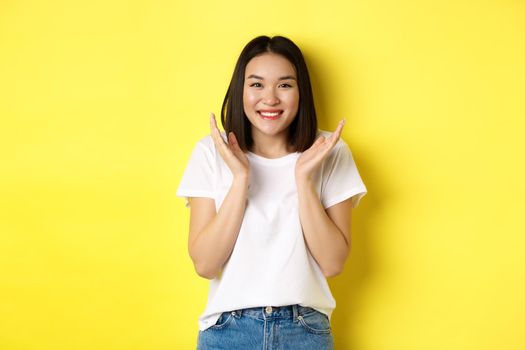 Beauty and fashion concept. Excited asian woman clap hands and smiling happy at camera, standing in white t-shirt against yellow background.