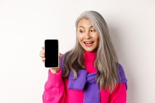 Online shopping. Stylish and beautiful asian senior woman with grey hair, showing blank smartphone screen and looking pleased, standing over white background.