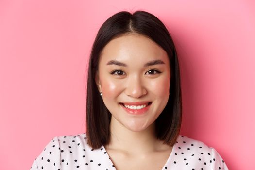 Beauty and lifestyle concept. Headshot of beautiful asian woman smiling, looking at camera happy and romantic, standing against pink background.