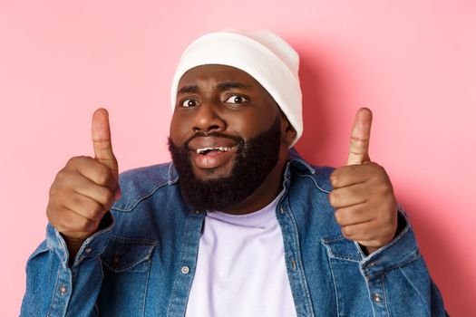 Close-up of awkward Black man in beanie showing thumbs-up but feeling cringe, standing unsure and worried against pink background.