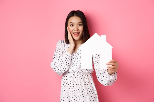 Real estate. Adult asian woman searching for home, holding house model and smiling, promo of broker company, standing over pink background.