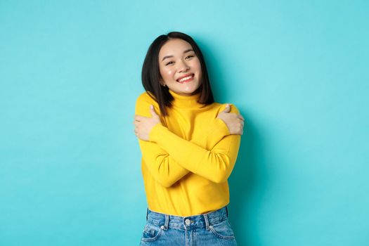 Cheerful beautiful girl embracing own body, smiling and cuddling herself, standing carefree in yellow pullover against blue background.