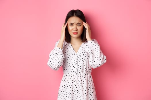 Image of upset asian woman having headache, feeling unwell or dizzy, close eyes and massaging head, suffering migraine, standing over pink background.