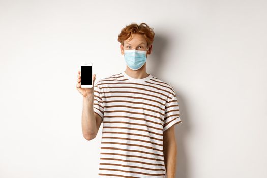 Covid-19, online doctor concept. Smiling redhead man in face mask showing smartphone screen, standing over white background.