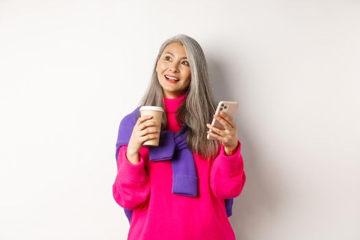 Fashionable asian mature woman with grey hair, drinking coffee and using smartphone, looking upper left corner promotion with dreamy face, white background.