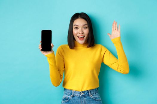 Cheerful asian woman showing empty smartphone screen and raising hand to say hello, greeting you, standing against blue background.