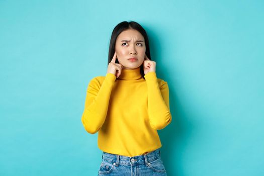 Annoyed korean girl complaining on loud neighbours, shut ears with fingers and looking angry up, standing against blue background.