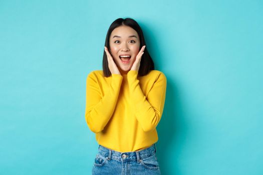 Portrait of pretty korean girl receive surprising news, looking amazed and happy at camera, standing over blue background.