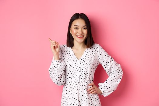 Cute asian woman in dress pointing upper left corner copy space. Korean model showing advertisement, standing over pink background.