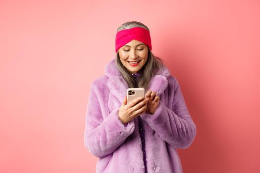 Online shopping and fashion concept. Modern asian granny in trendy faux fur coat texting message, using smartphone and looking happy at screen, pink background.