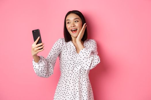 Beautiful asian girl using photo filters app and taking selfie on smartphone, posing in cute dress against pink background.