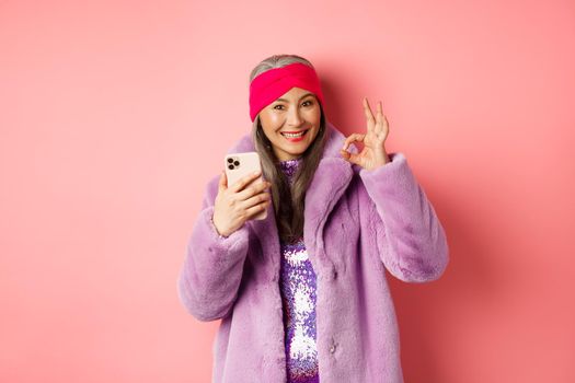 Online shopping and fashion concept. Stylish asian senior woman showing okay sign and holding mobile phone, recommending internet store, pink background.