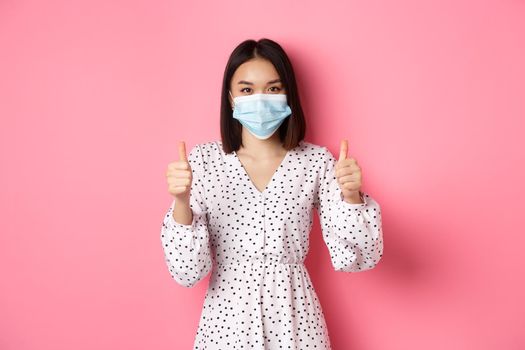 Covid-19, quarantine and lifestyle concept. Beautiful asian woman in dress and face mask showing thumbs-up, give approval, like and praise, standing over pink background.