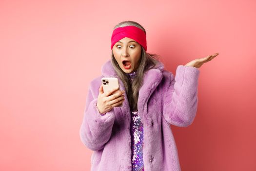 Online shopping and fashion concept. Amazed and surprised asian senior woman looking at smartphone screen, reading on mobile phone and gasping, pink background.