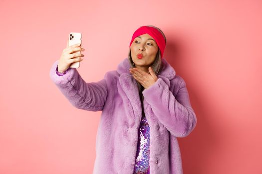 Fashion concept. Beautiful and trendy asian mature woman in faux fur coat with party dress, taking selfie, sending kiss to smartphone camera, posing against pink background.
