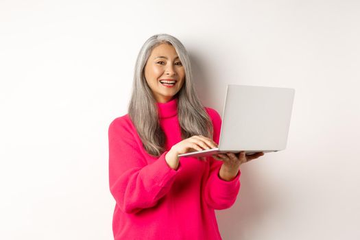 Beautiful asian senior woman entrepreneur working with laptop, laughing and smiling at camera, standing over white background.