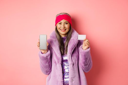 Online shopping and fashion concept. Fashionable asian senior woman showing black screen of smartphone and plastic credit card, smiling happy, pink background.