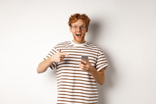 Technology and e-commerce concept. Amazed redhead man checking out online promo offer on mobile phone, pointing finger at smartphone and smiling, white background.