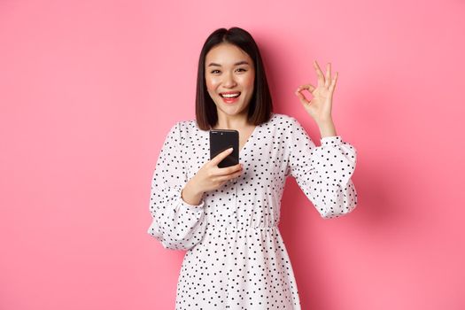 Online shopping and beauty concept. Satisfied asian female customer showing okay, making purchase in internet on smartphone, standing over pink background.