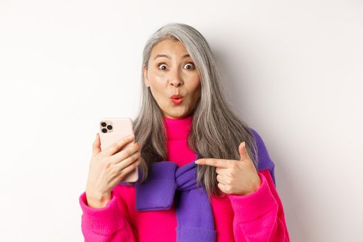 Online shopping. Close up of amazed asian senior woman pointing at smartphone and smiling amazed, standing over white background.