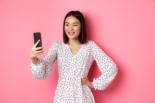 Beautiful asian girl using photo filters app and taking selfie on smartphone, posing in cute dress against pink background.