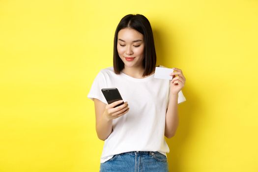 E-commerce and online shopping concept. Beautiful asian woman paying in internet, looking at smartphone screen and holding plastic credit card, yellow background.