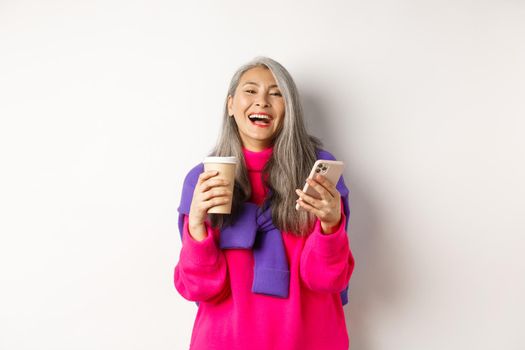 Social networking. Happy asian senior woman drinking coffee and holding smartphone, laughing at camera, standing over white background.