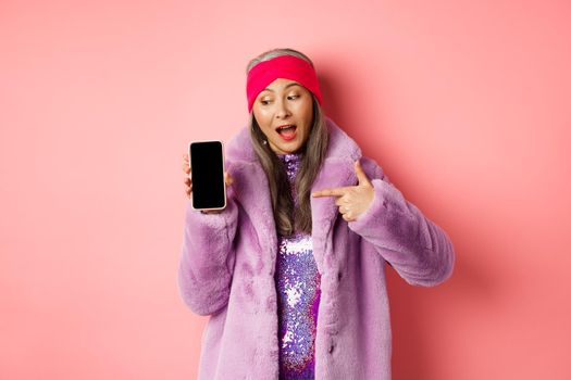 Online shopping and fashion concept. Cool asian granny in stylish faux fur coat pointing finger left at blank smartphone screen, checking out internet promo, pink background.