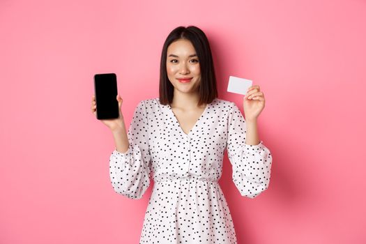 Cute asian woman shopping online, showing bank credit card and mobile screen, smiling and looking at camera, standing over pink background.