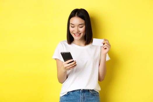 E-commerce and online shopping concept. Beautiful asian woman paying in internet, looking at smartphone screen and holding plastic credit card, yellow background.