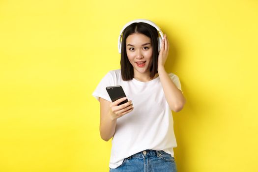 Modern asian woman listening music in wireless headphones, reading smartphone screen and smiling, standing in white t-shirt over yellow background.