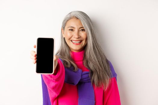 Online shopping. Close up of modern asian senior woman extending hand with mobile phone, showing blank smartphone screen and smiling, standing over white background.